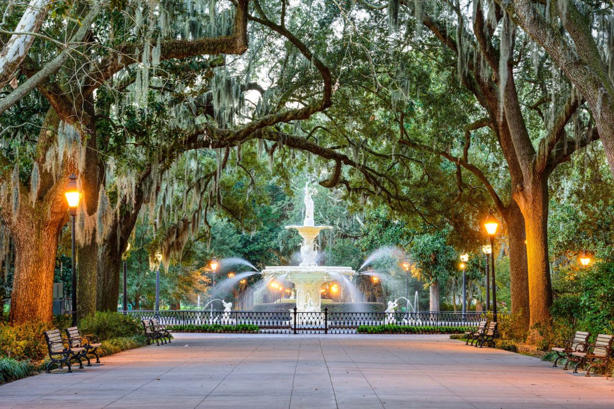 Evening view of a decorative fountain in a moss-draped, tree-lined park with benches and lampposts. (centered fountain, large trees dripping with Spanish moss)